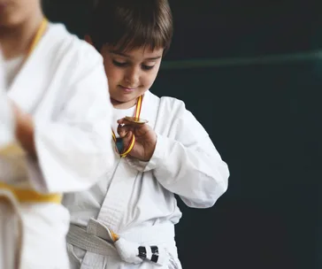 A young child in a martial arts uniform examines a medal around their neck, focused and proud.
