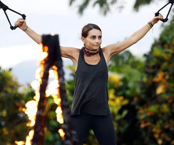 Pia Miranda in a challenge on Survivor, holding ropes with outstretched arms beside a fire.