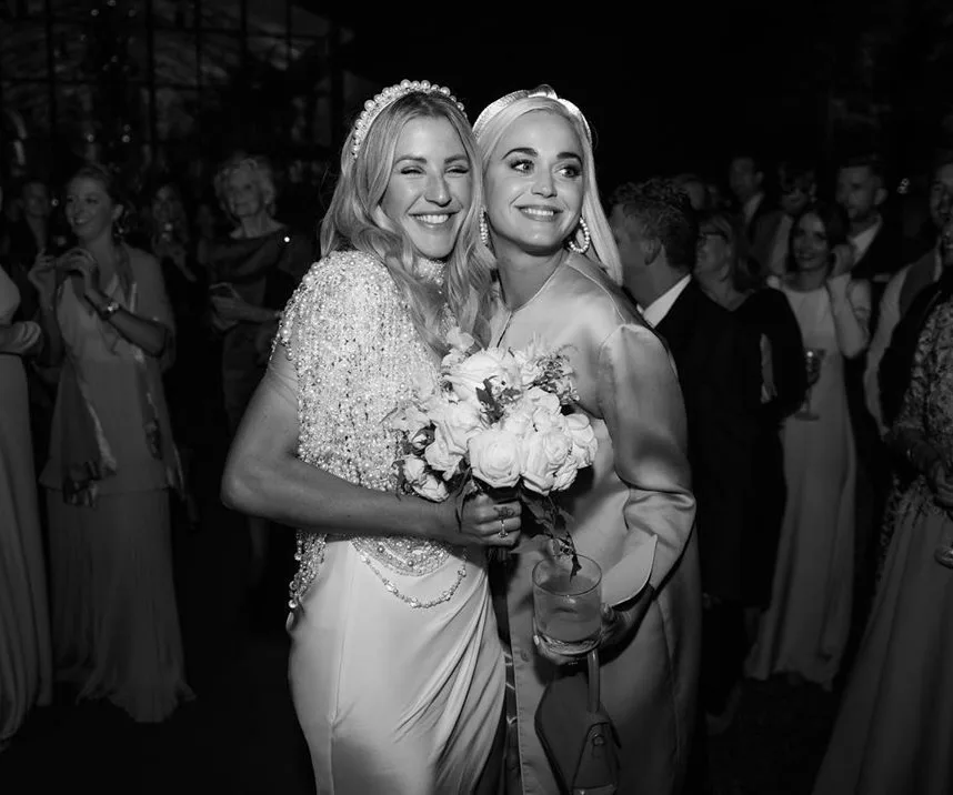 Bride and guest smiling at wedding, bride holding bouquet, surrounded by people.