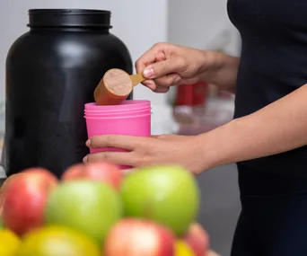Person scooping protein powder into a pink cup with fruits and large black container nearby.