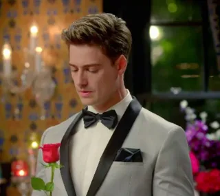 Man in a tuxedo holding a single red rose, standing in a dimly lit, decorated room.