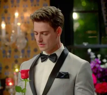 Man in a tuxedo holding a single red rose, standing in a dimly lit, decorated room.