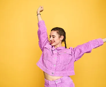 Person in purple corduroy outfit smiling with arms raised against a yellow background.