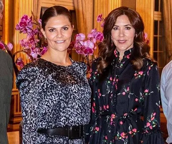 Two women stand together indoors, both smiling, with orchid flowers in the background.