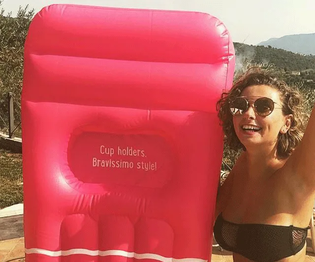 A woman in sunglasses poses with a large pink inflatable pool lounger with the text "Cup holders. Bravissimo style!"