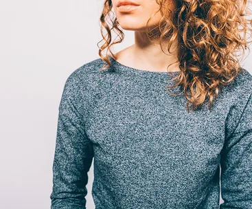 Curly-haired woman in a grey sweater showing split ends, focus on hair texture.