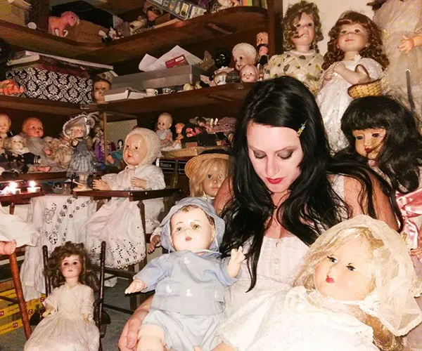 A woman sits among a collection of vintage dolls in a dimly lit room, creating a spooky atmosphere.