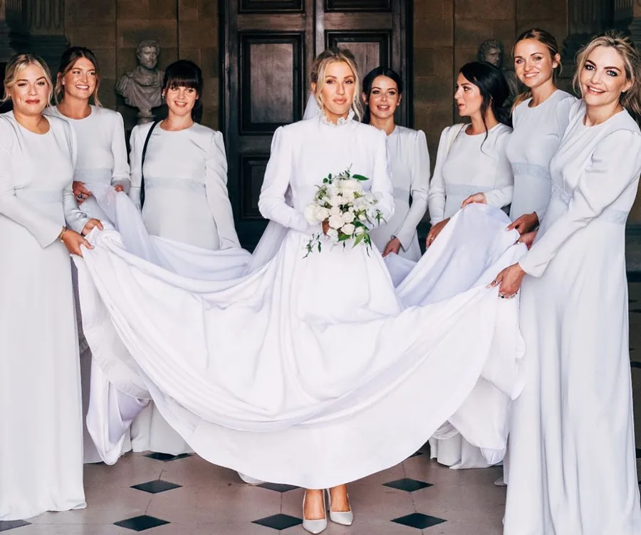 Bride in a white gown holding a bouquet, surrounded by seven bridesmaids in matching white dresses.