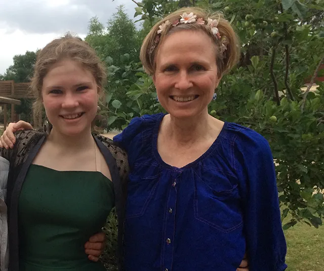 Two people smiling, standing outside near green trees. One wears a dark green dress, the other a blue top with a floral headband.
