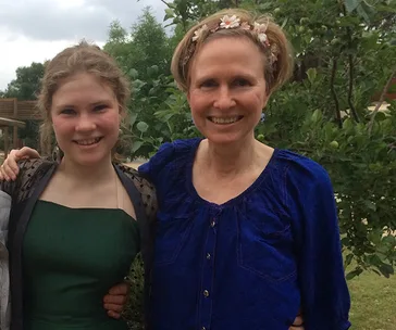 Two people smiling, standing outside near green trees. One wears a dark green dress, the other a blue top with a floral headband.