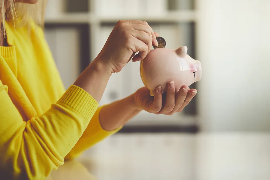 Person in yellow sweater placing coin in a pink piggy bank on a light-colored surface.