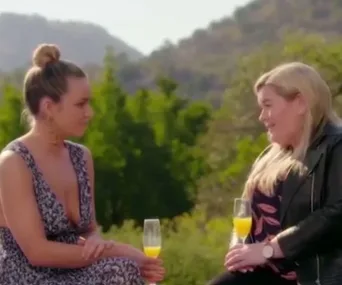 Two women seated outside with drinks, engaging in conversation against a backdrop of greenery and mountains.