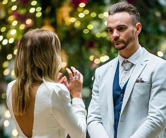 A woman in a white dress talks to a man in a light suit with bokeh lights in the background, from "The Bachelorette."