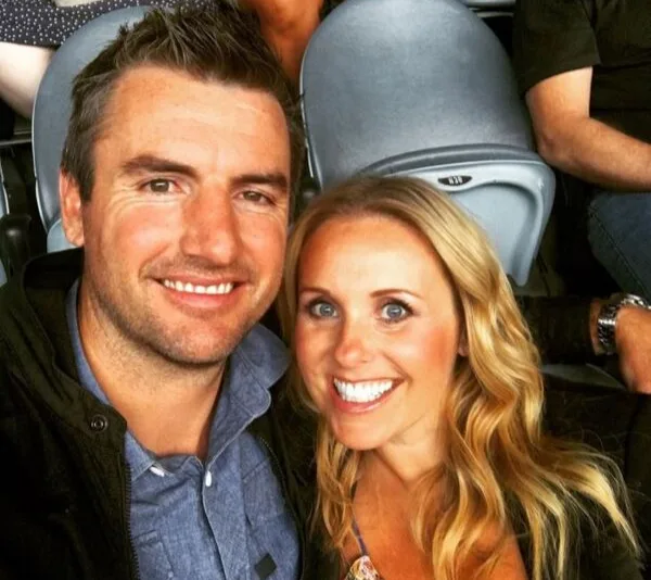 A smiling couple taking a selfie in a stadium with chairs in the background.