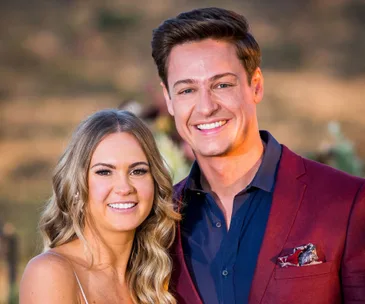 A couple smiling outdoors; man in maroon blazer, woman with long blonde hair, blurred natural background.