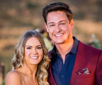 A couple smiling outdoors; man in maroon blazer, woman with long blonde hair, blurred natural background.