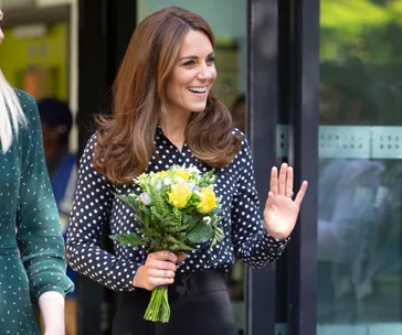 A woman in a polka dot top holds flowers and waves while smiling outside a building.