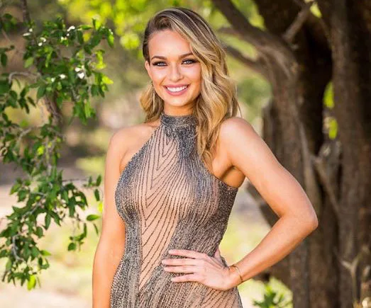 A woman in an elegant dress stands outdoors, smiling with her hand on her hip, surrounded by trees.