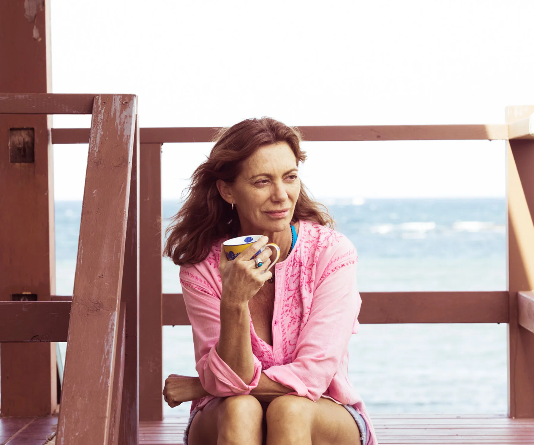 A woman in a pink shirt sits on a wooden deck, holding a mug, with the ocean in the background.