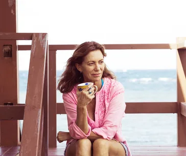 A woman in a pink shirt sits on a wooden deck, holding a mug, with the ocean in the background.