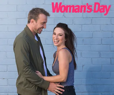 Man and woman laughing together in front of blue brick wall with "Woman's Day" logo.