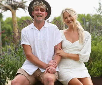 A couple sitting outdoors, the man in a white shirt and hat, the woman in a white dress, both smiling at the camera.