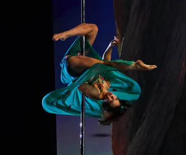 Pole dancer in a teal costume performs an inverted pose on stage during a dynamic performance.