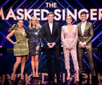 The Masked Singer judges and host in elegant outfits on stage, with show logo in the background.