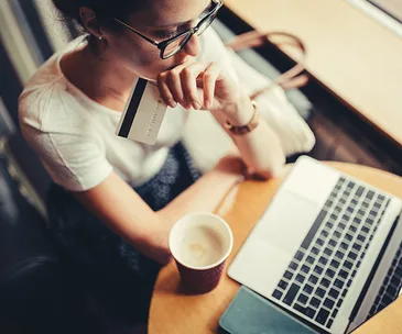 Woman with a credit card looking at a laptop, seated at a café table with a cup of coffee.