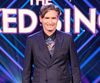 Dave Hughes, smiling in a dark suit, stands in front of "The Masked Singer" sign with a colorful background.