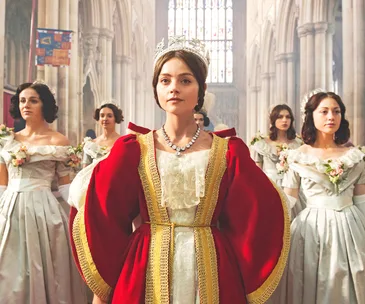 Woman in royal attire and crown with bridesmaids in a grand hall.