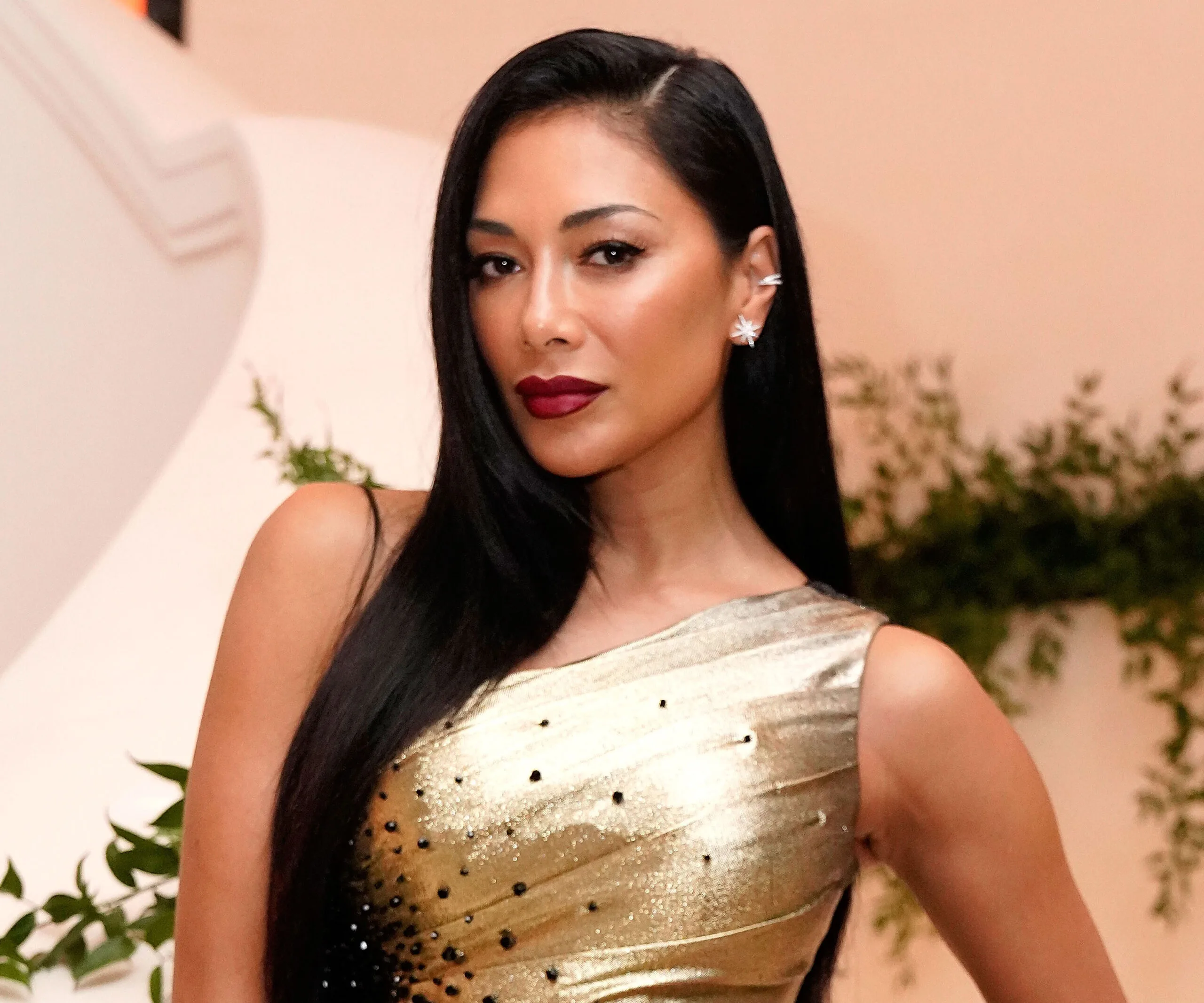 Nicole Scherzinger in a metallic gold dress with long black hair, posing elegantly indoors.