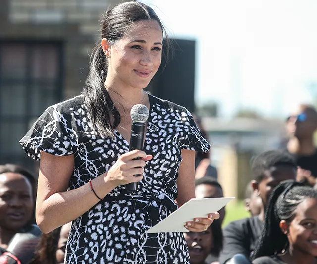 "Meghan Markle speaking outdoors with microphone and notes, audience in background."