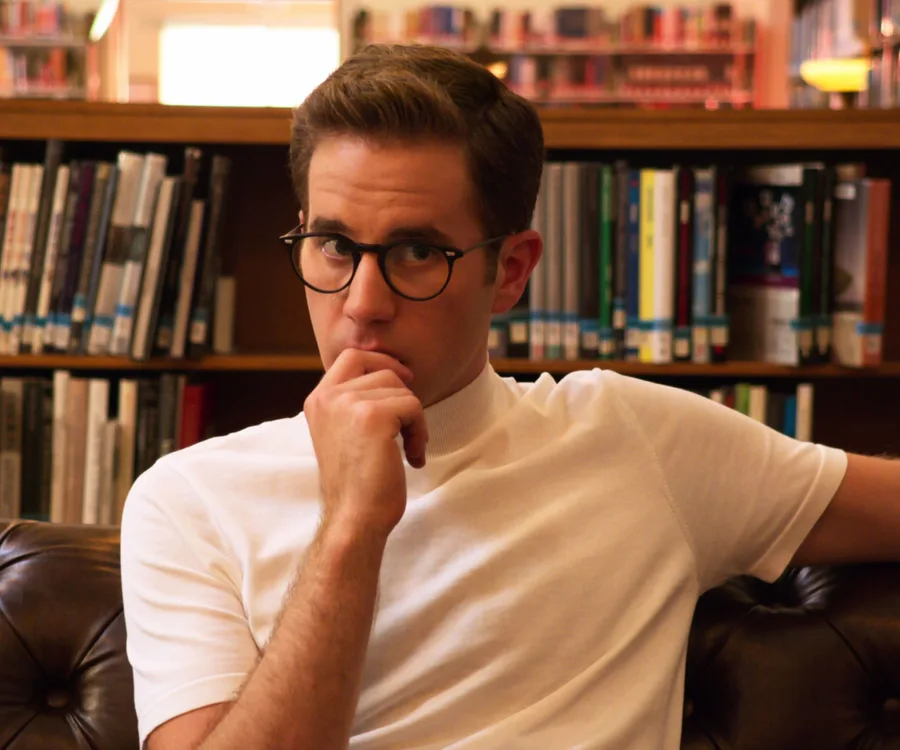 A person with glasses in a library, seated on a brown leather couch, looking thoughtfully at the camera.