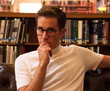 A person with glasses in a library, seated on a brown leather couch, looking thoughtfully at the camera.