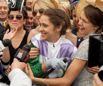 A smiling jockey in colorful silks is hugged by jubilant fans while surrounded by microphones.