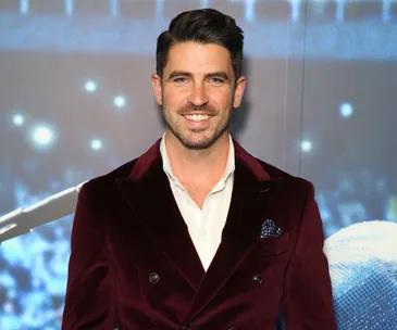 A man in a dark red blazer stands smiling against a blurred blue-lit background.