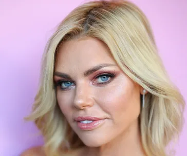 Blonde woman with styled hair and makeup against a pink background.