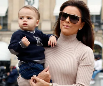 Woman in sunglasses holds a young child outdoors, both wearing stylish clothes.