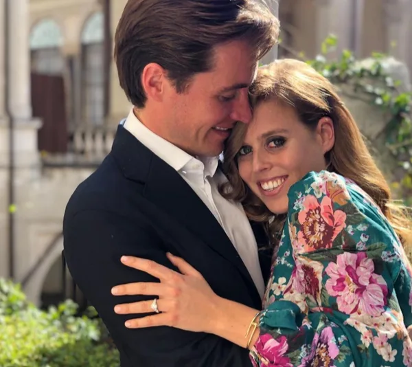 A couple embracing, the woman wearing a floral dress and showing an engagement ring, smiling at the camera.