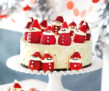 Cake topped with strawberry Santas, decorated with cream faces and hats, displayed on a white cake stand.