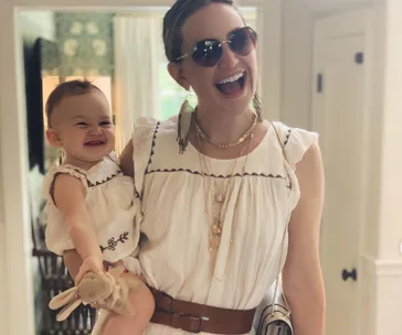 Mother and daughter in matching white dresses, both smiling, with the mother wearing sunglasses indoors.