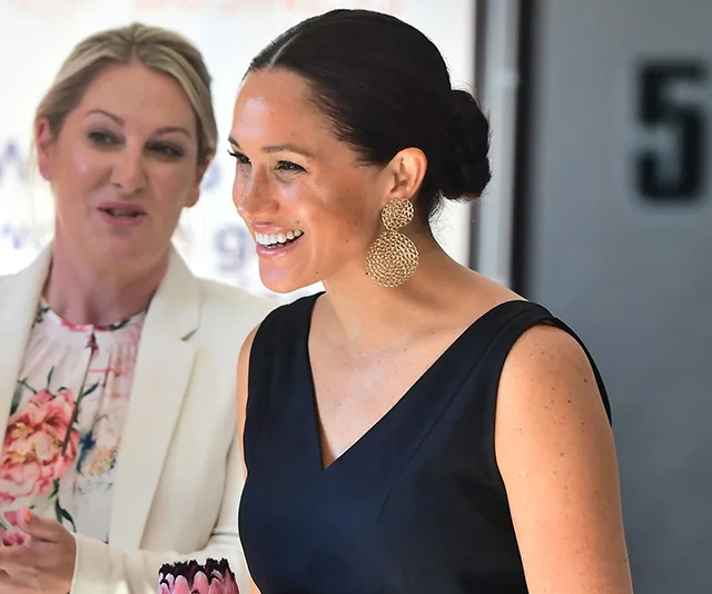 Meghan Markle in a black jumpsuit with large earrings, smiling and standing next to another woman.