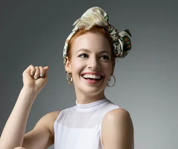 Smiling woman with a headscarf and hoop earrings, wearing a sleeveless white top against a gray background.