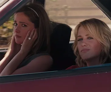 Two women sitting in a car, one is covering her ears looking uncomfortable, while the other is smiling slightly.