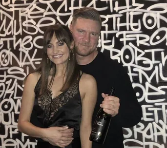 A couple posing together, smiling in front of a graffiti-style background, with the man holding a bottle.