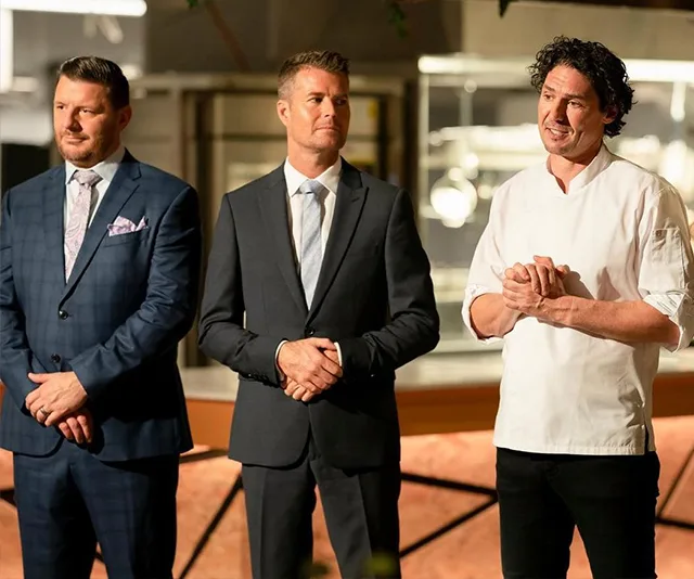 Three men in formal attire standing in a kitchen setting, serious expressions, likely on a cooking show.