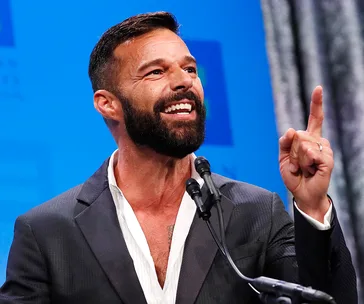 Man in a suit passionately speaking at a microphone with a raised finger gesture.