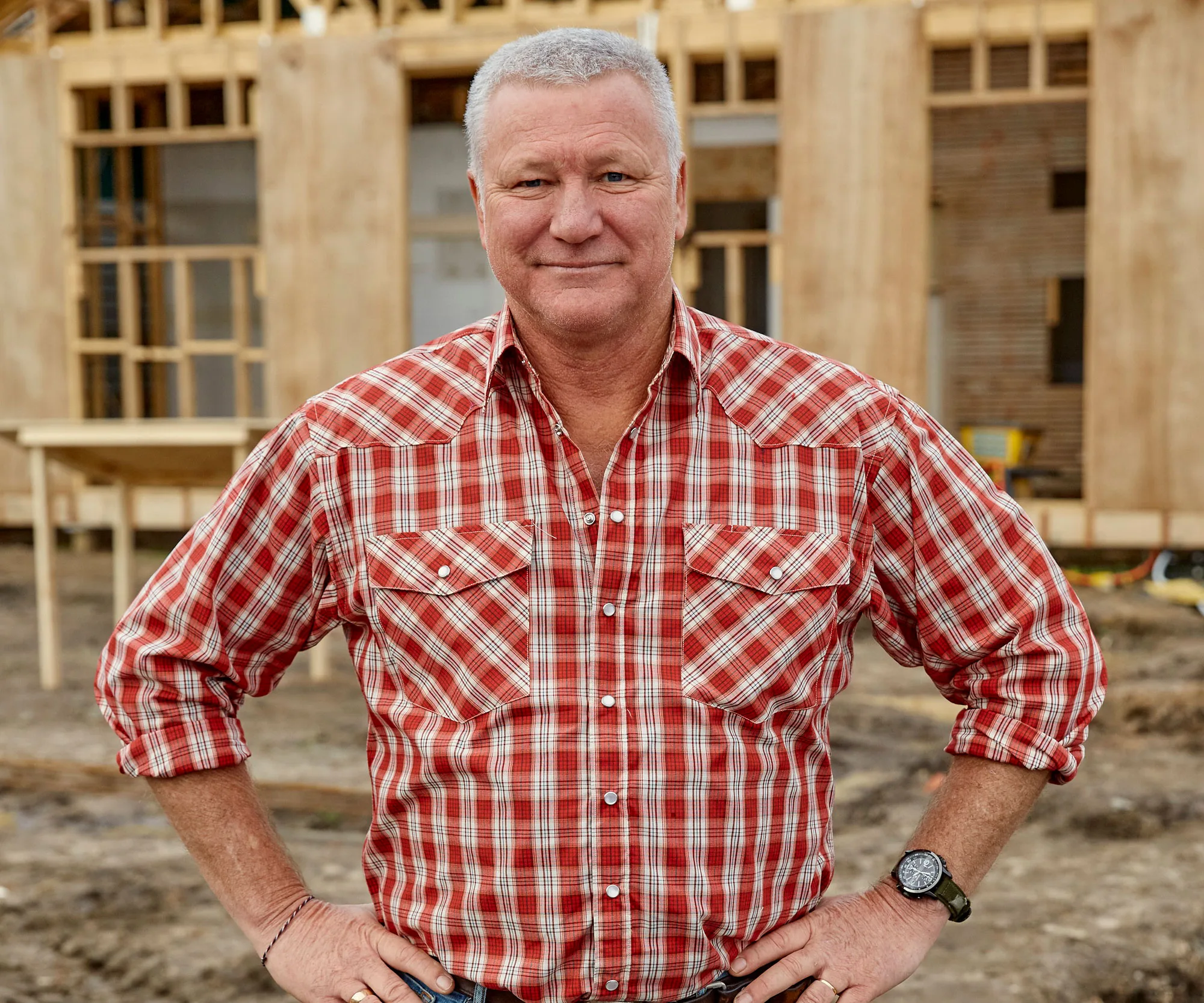 Man in a red plaid shirt standing at a construction site, wooden frame structure in the background.