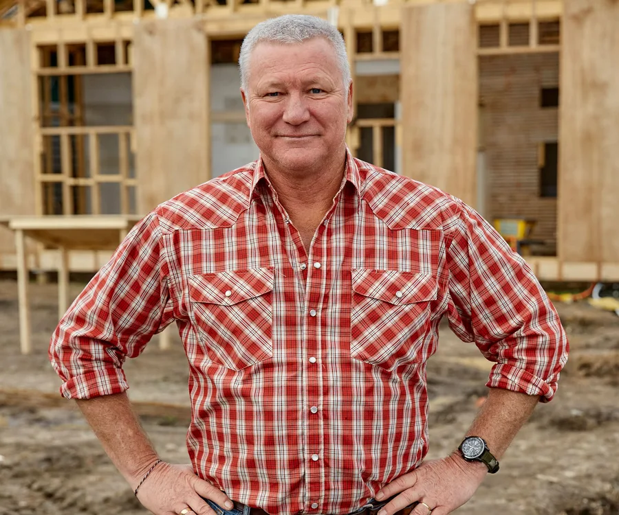 Man in a red plaid shirt standing at a construction site, wooden frame structure in the background.